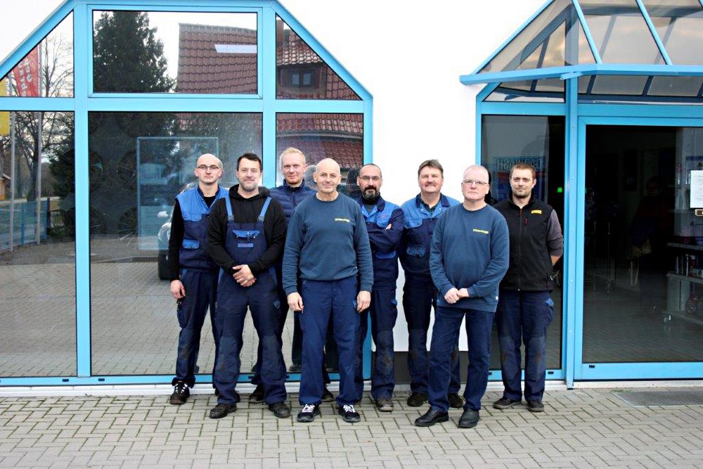 Das Bild zeigt Mitarbeiter in Arbeitskleidung, die vor einem Gebäude mit großen blauen Fenstern stehen.