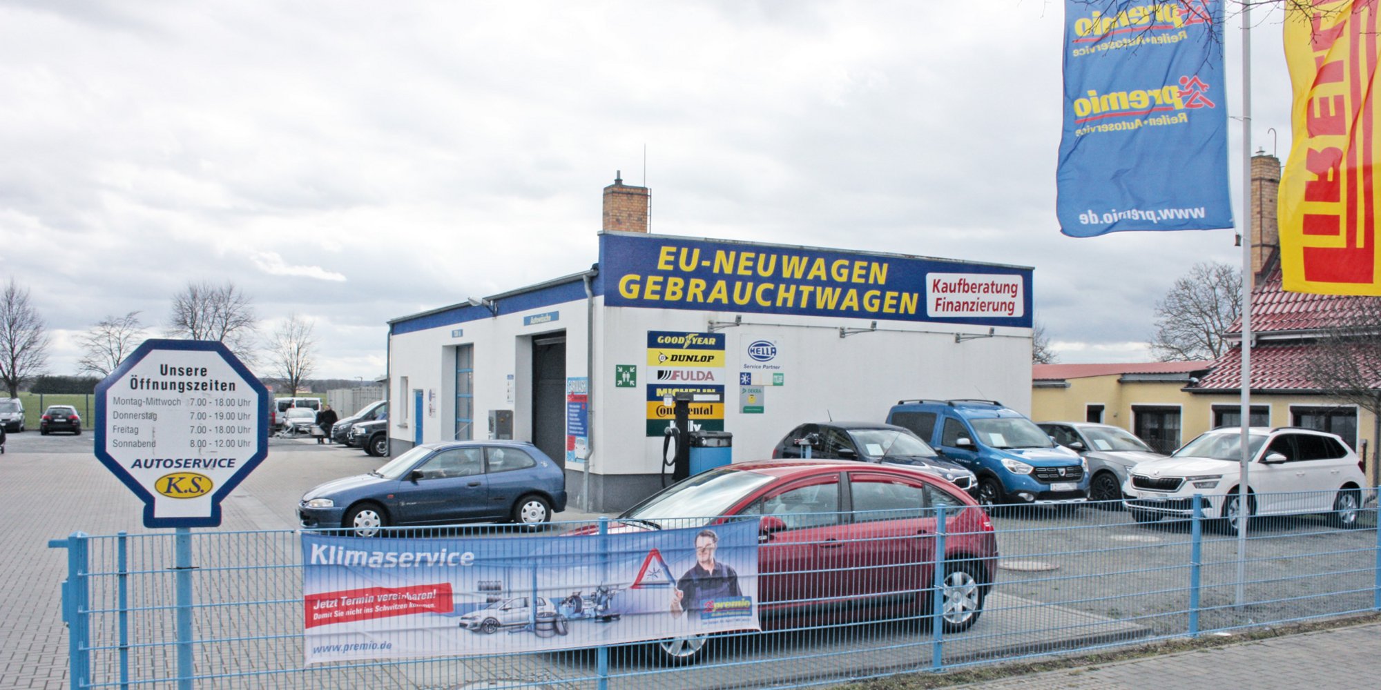 Das Bild zeigt eine Werkstatt mit Parkplatz, mehreren Autos, Fahnen und Schildern. Im Hintergrund sind Bäume und Gebäude zu sehen.